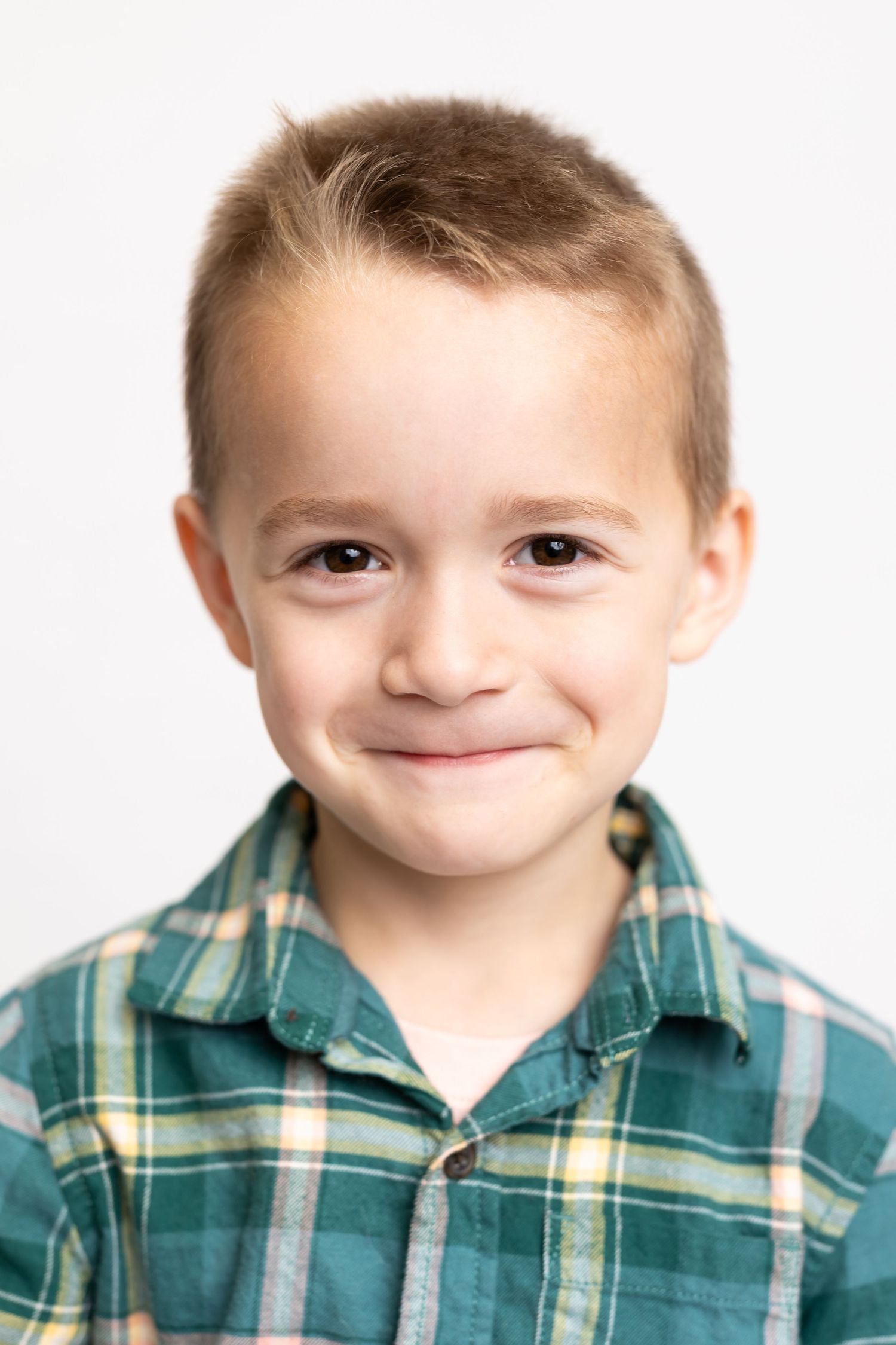 Child wearing plaid green and white shirt smiling softly against white backdrop.