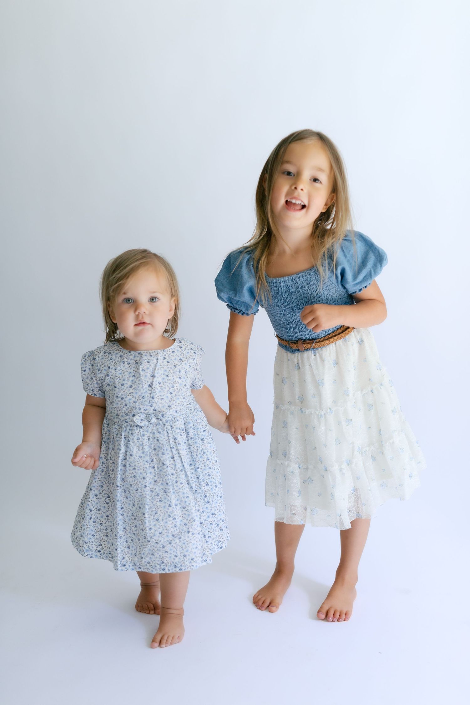 Two young girls in floral and denim dresses holding hands, smiling and posing barefoot on white background.