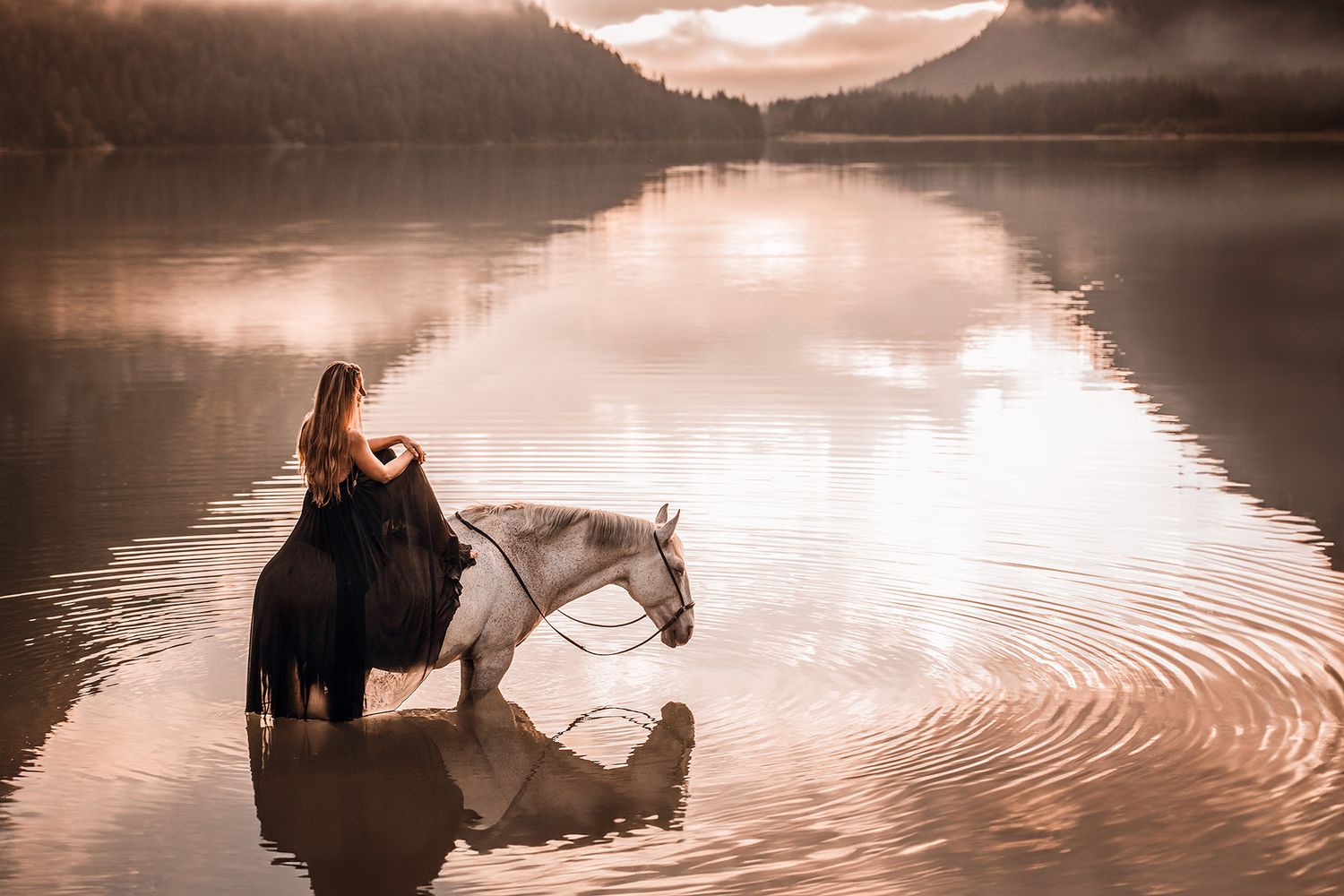 Frau in schwarzem Kleid sitzt auf weißem Pferd, das im Wasser mit Bergpanorama steht.