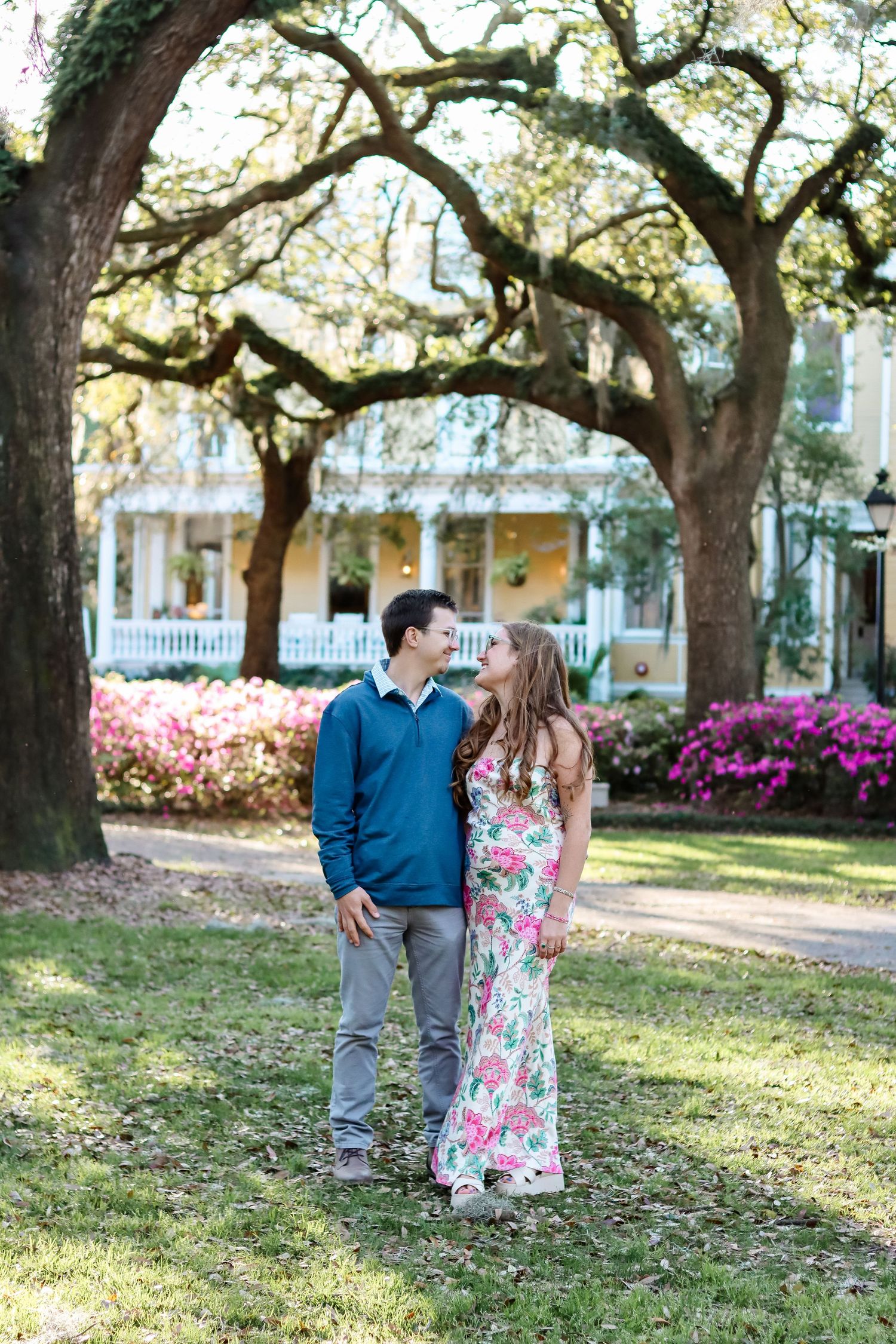 Couple shares romantic moment under moss-draped oak trees with pink azaleas blooming in background.