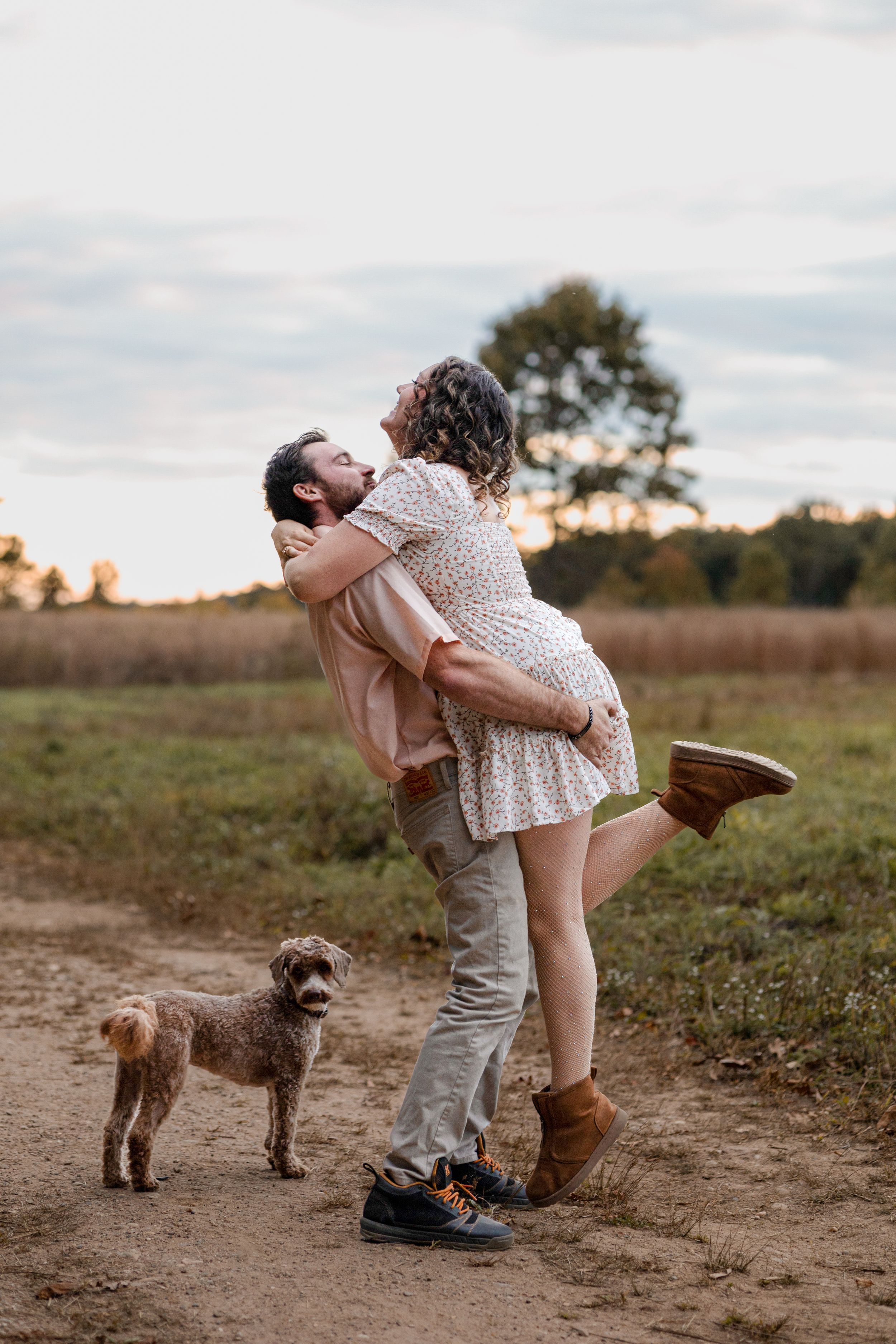Romantic couple embraces in a field at sunset while their small dog watches nearby.
