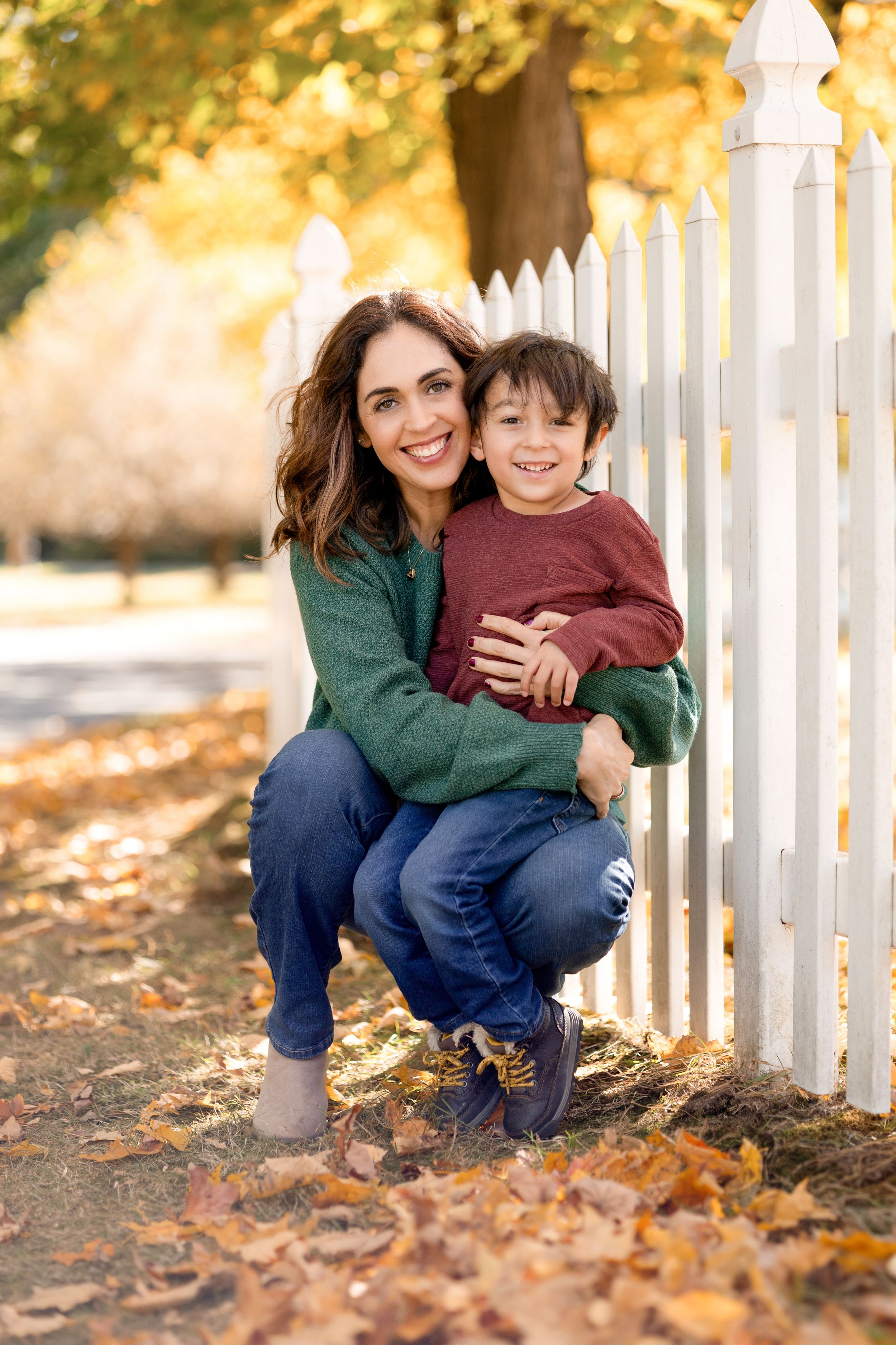 Parent and child share playful moments by white picket fence surrounded by autumn leaves.