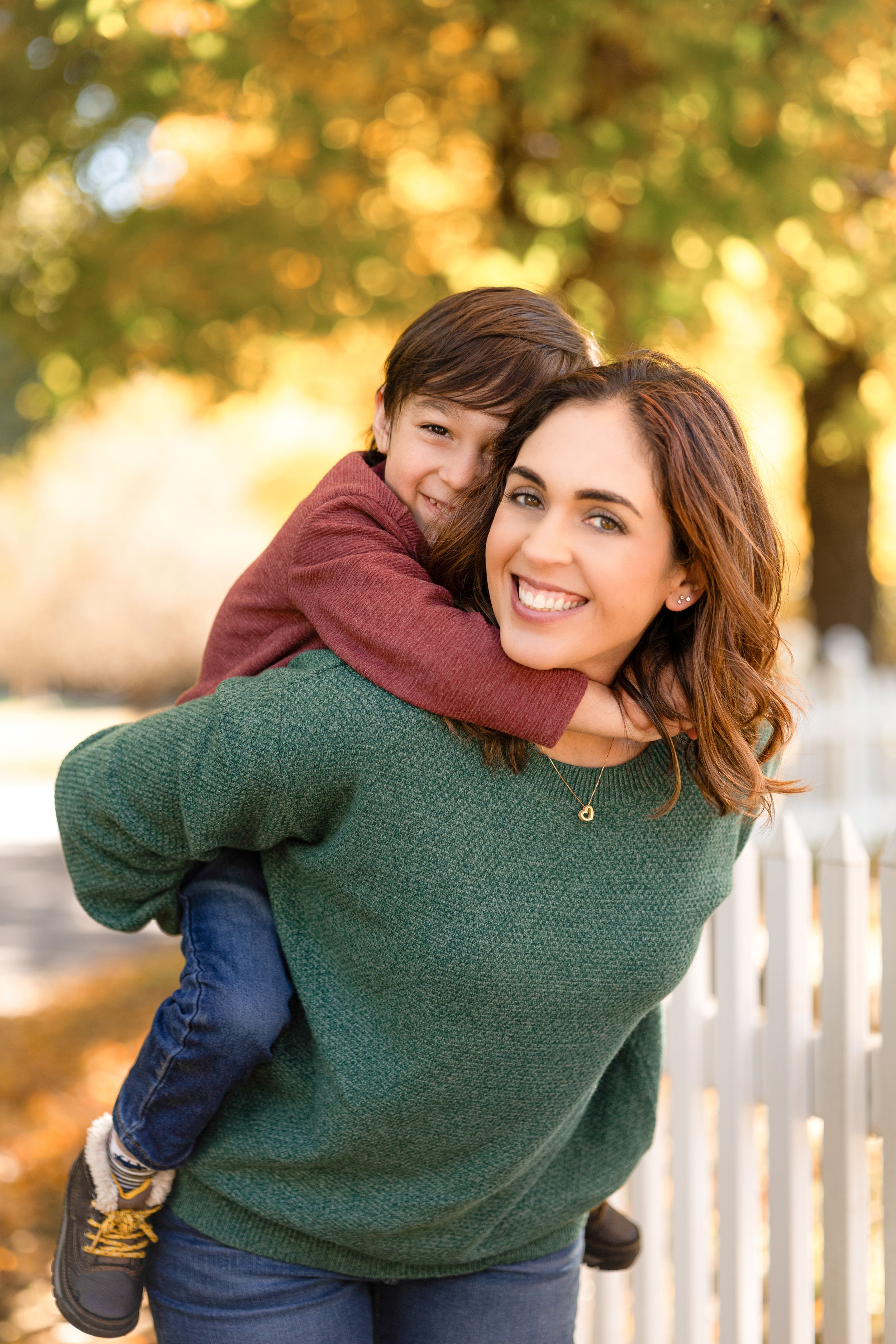 Parent gives child piggyback ride along white picket fence in fall setting.