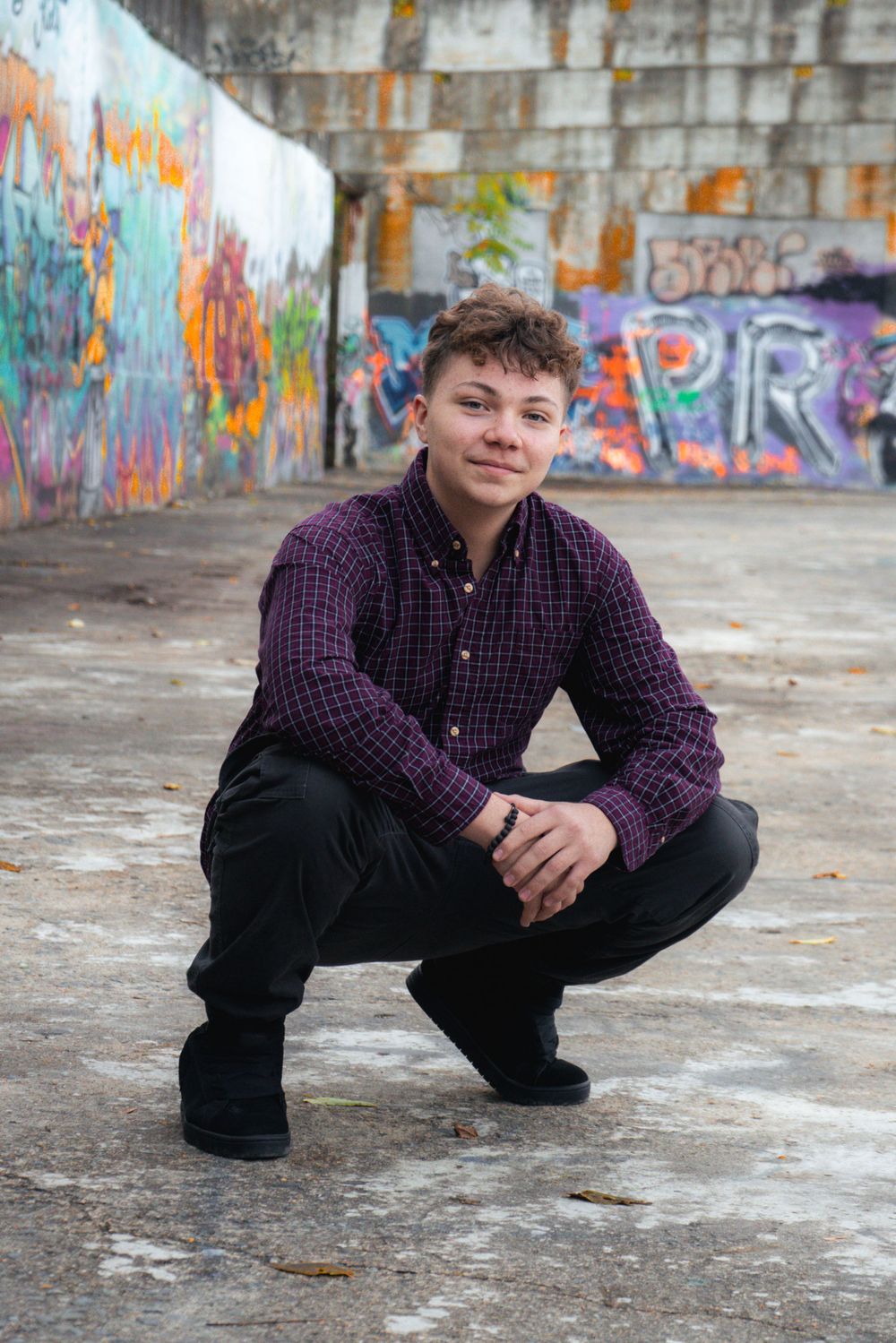 Portrait against colorful graffiti wall wearing purple plaid shirt and black pants in urban setting.