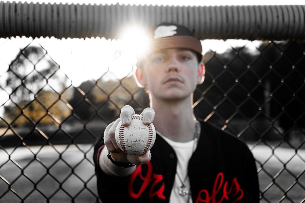 Baseball player holding ball against chain link fence with dramatic lighting.