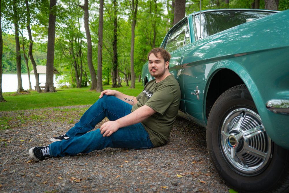 A person relaxes near a parked vintage turquoise car in a wooded area on a cloudy day.