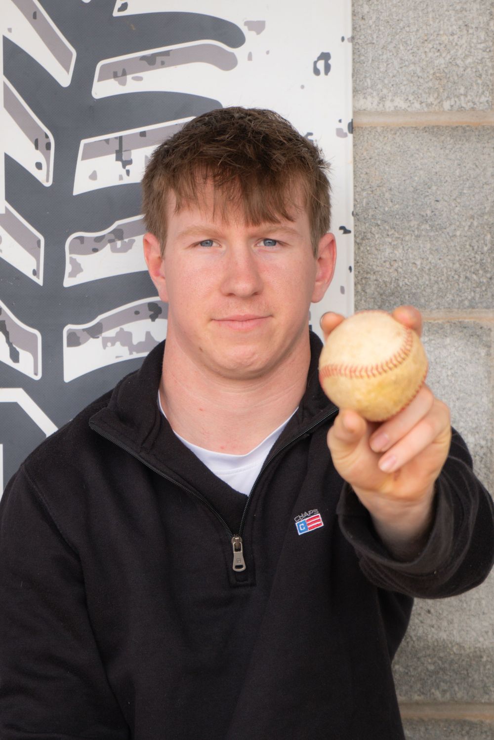 A person in a black zip-up jacket holds up a baseball while standing against a wall with graphic elements.