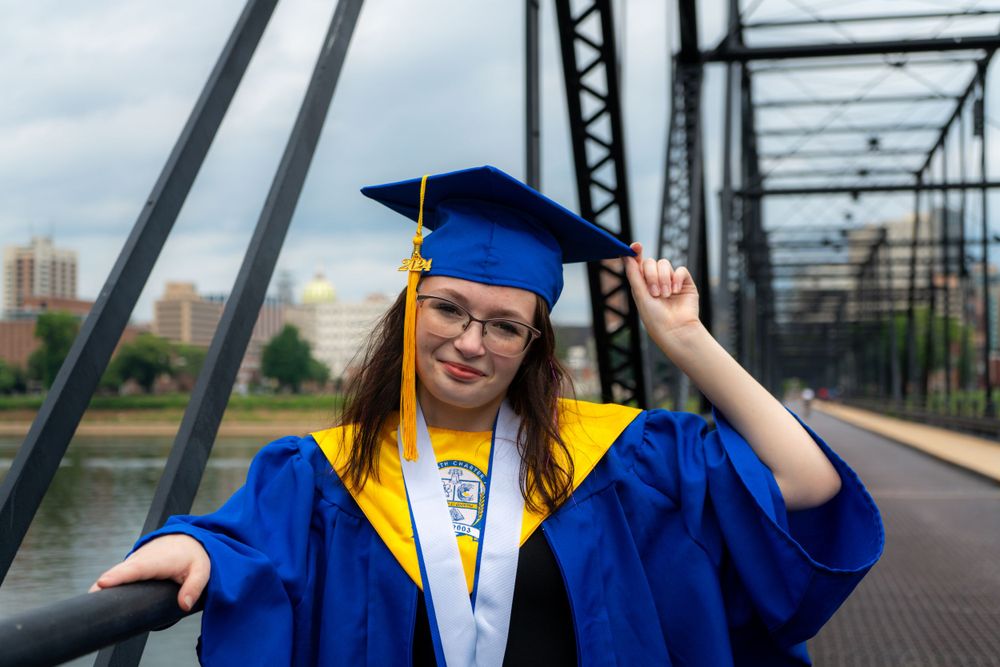 Graduate in blue and gold regalia poses on bridge with city skyline in background.