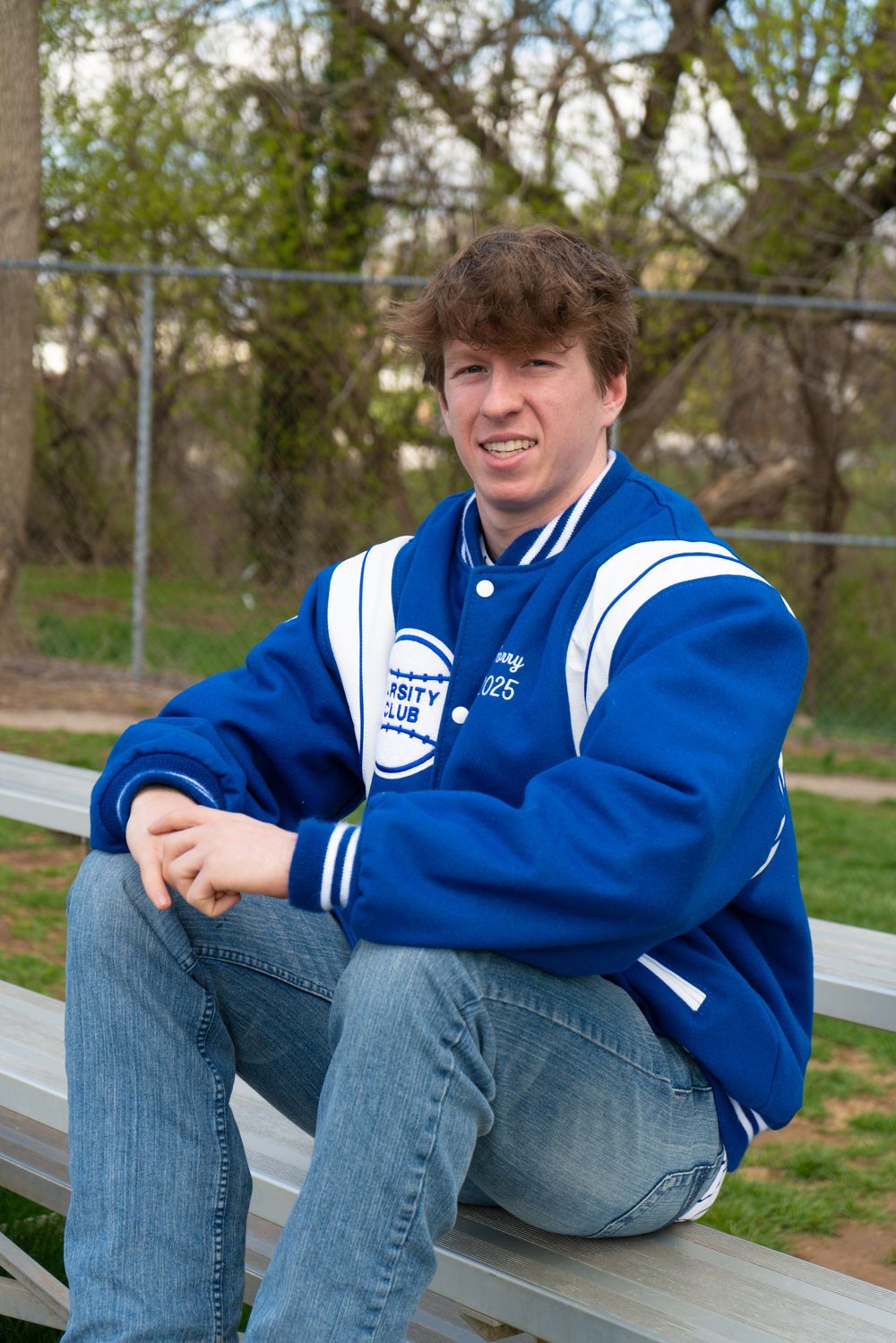 A person wearing a blue and white varsity letterman jacket sits casually on metal bleachers outdoors at a park.