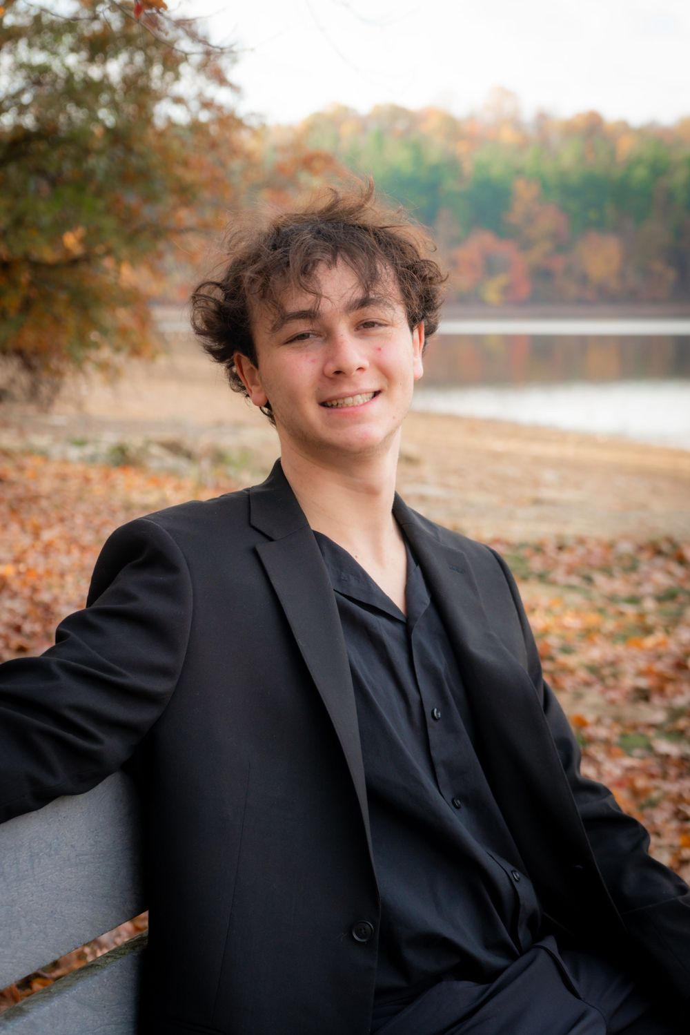 Portrait of a person in all black attire sitting outdoors during autumn with fall foliage and a lake in the background.