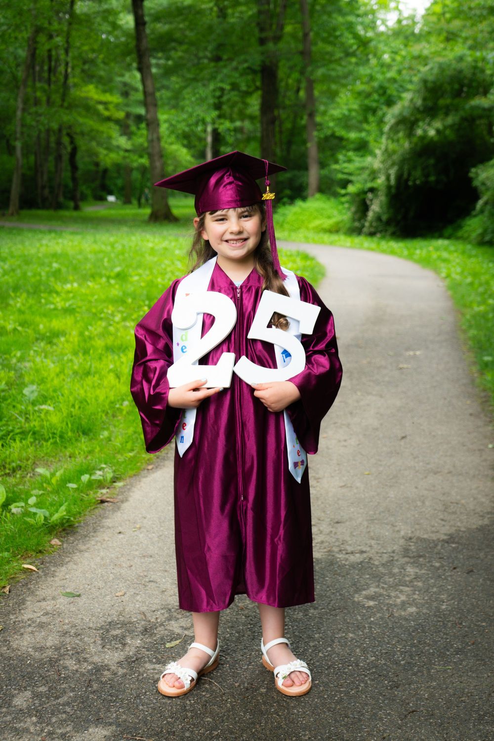 Graduate in maroon cap and gown holds decorative letters while standing on path in lush green forest setting.