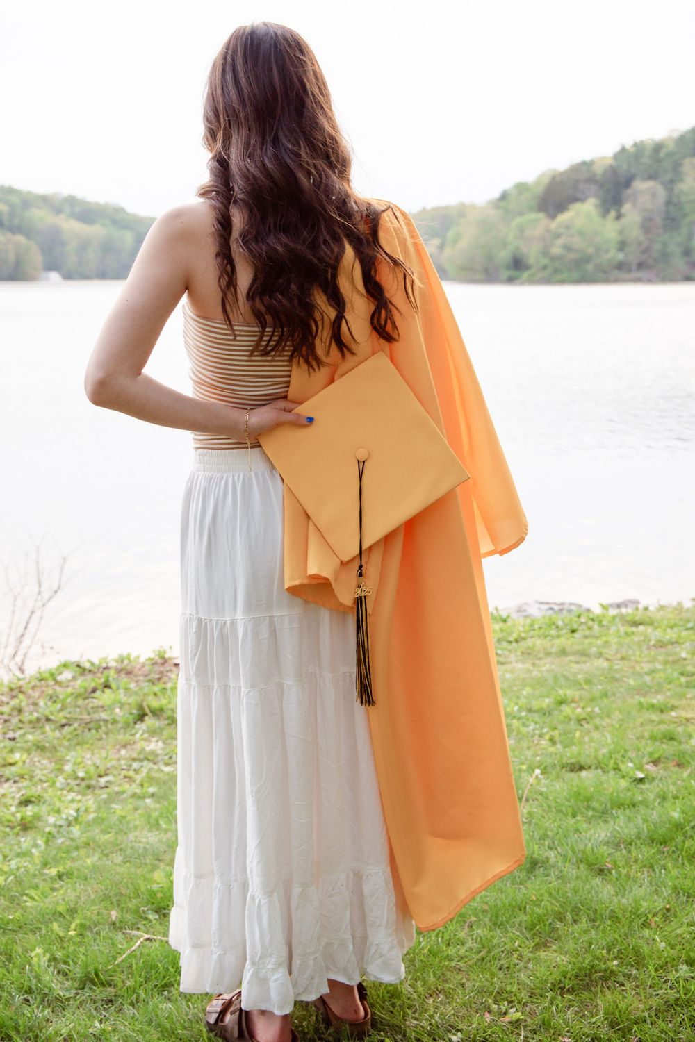 Graduate in white dress holds orange graduation gown and cap while standing by serene lake on grassy shore.