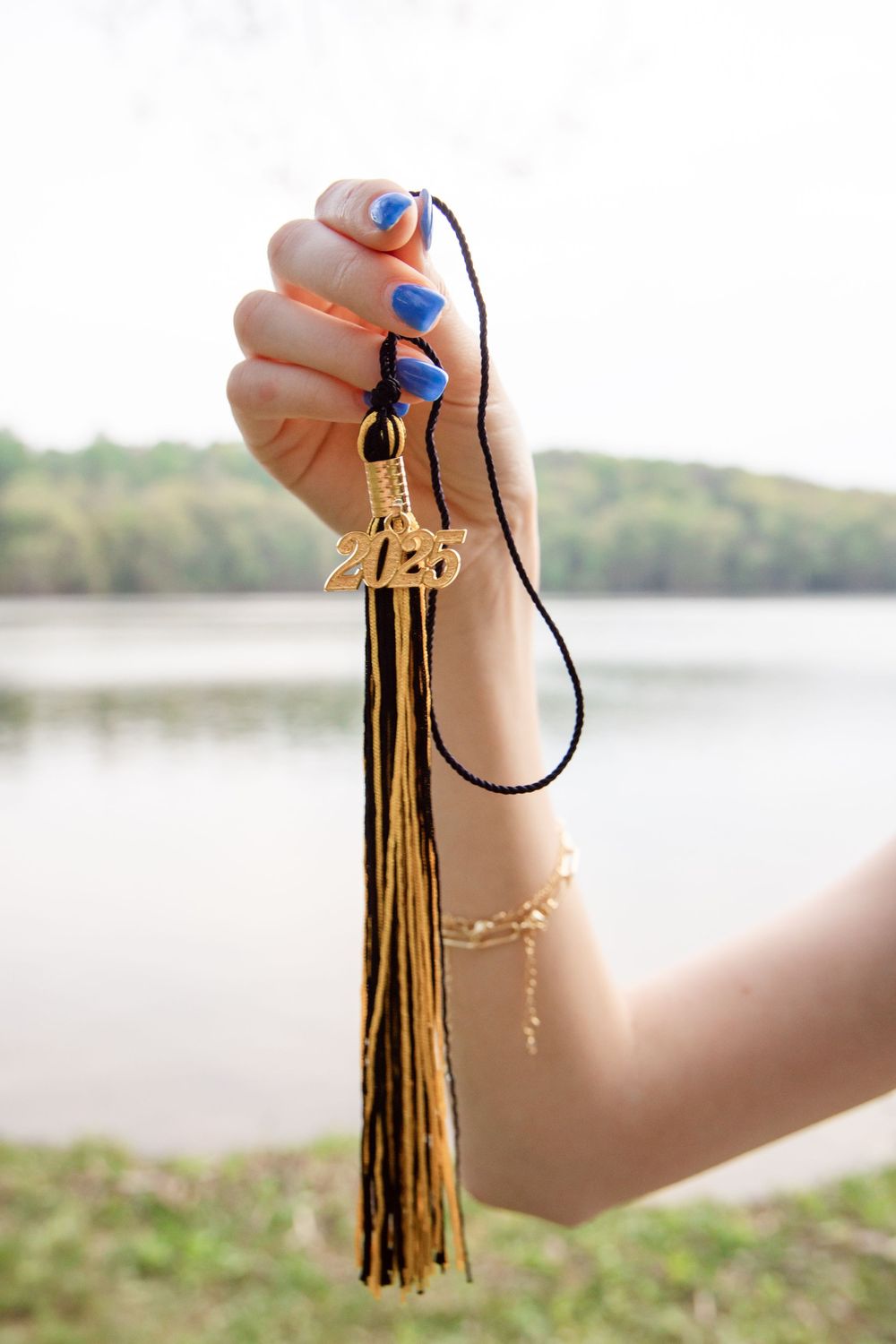 Hand with blue nail polish holds a brown leather tassel keychain against a serene lakeside background.