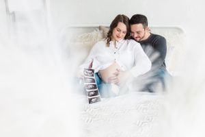Dreamy maternity photo of couple with ultrasound strip on white bed.