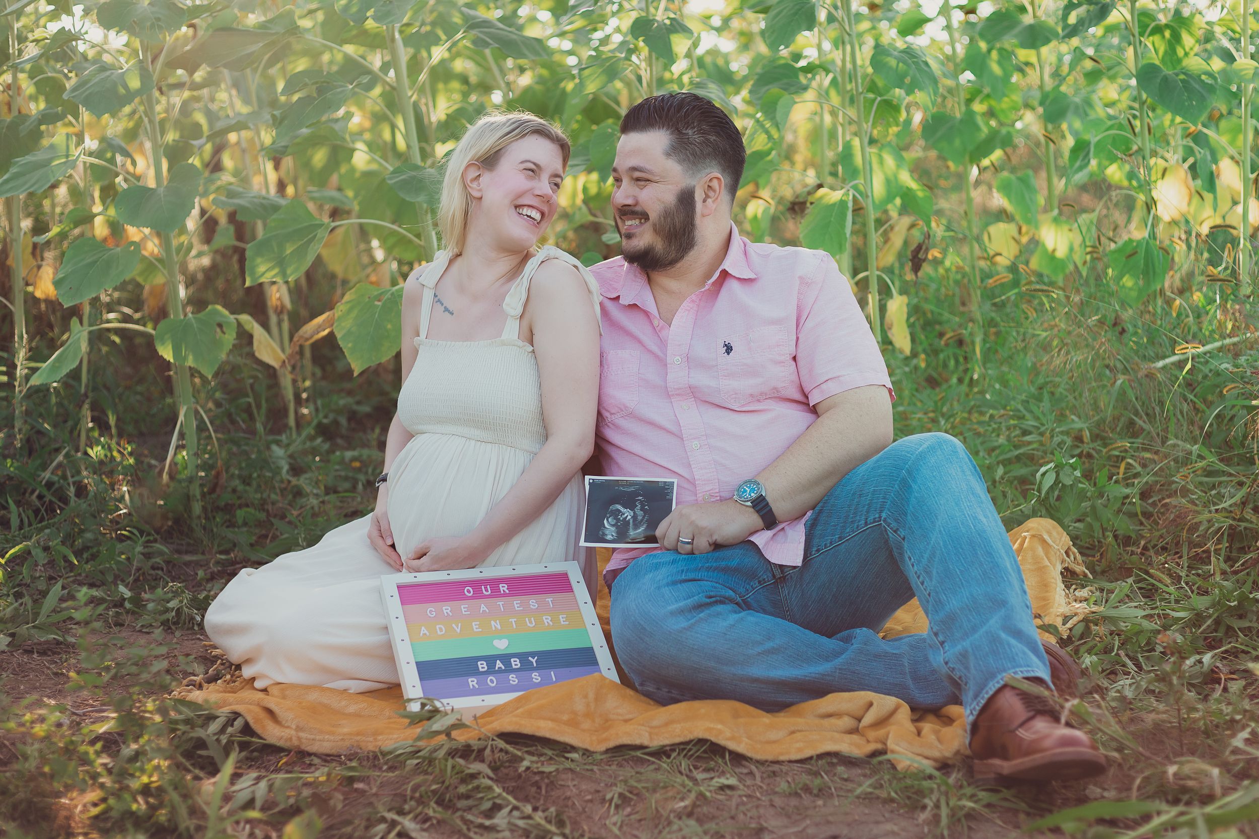 A couple sits on a blanket outdoors holding a rainbow-colored sign and ultrasound photo.