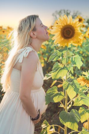 A woman in a white dress poses next to a large sunflower in a field at sunset.