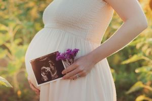 A close-up of hands holding an ultrasound photo with purple flowers against a white dress.