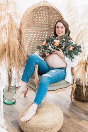 A maternity photo featuring a peacock chair, floral bouquet, and boho decor.