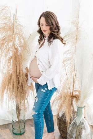 Expecting mother in white button down shirt and distressed jeans poses against pampas grass backdrop.
