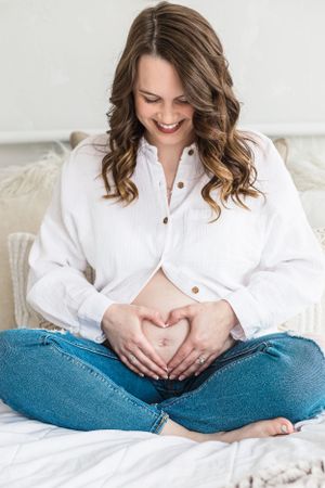 Expectant mother in white shirt and jeans makes heart shape with hands on belly.