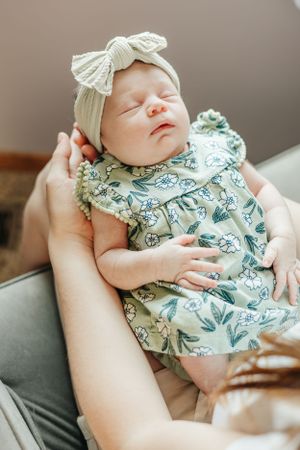 Peaceful newborn wearing cream bow and floral outfit sleeps on gray couch.