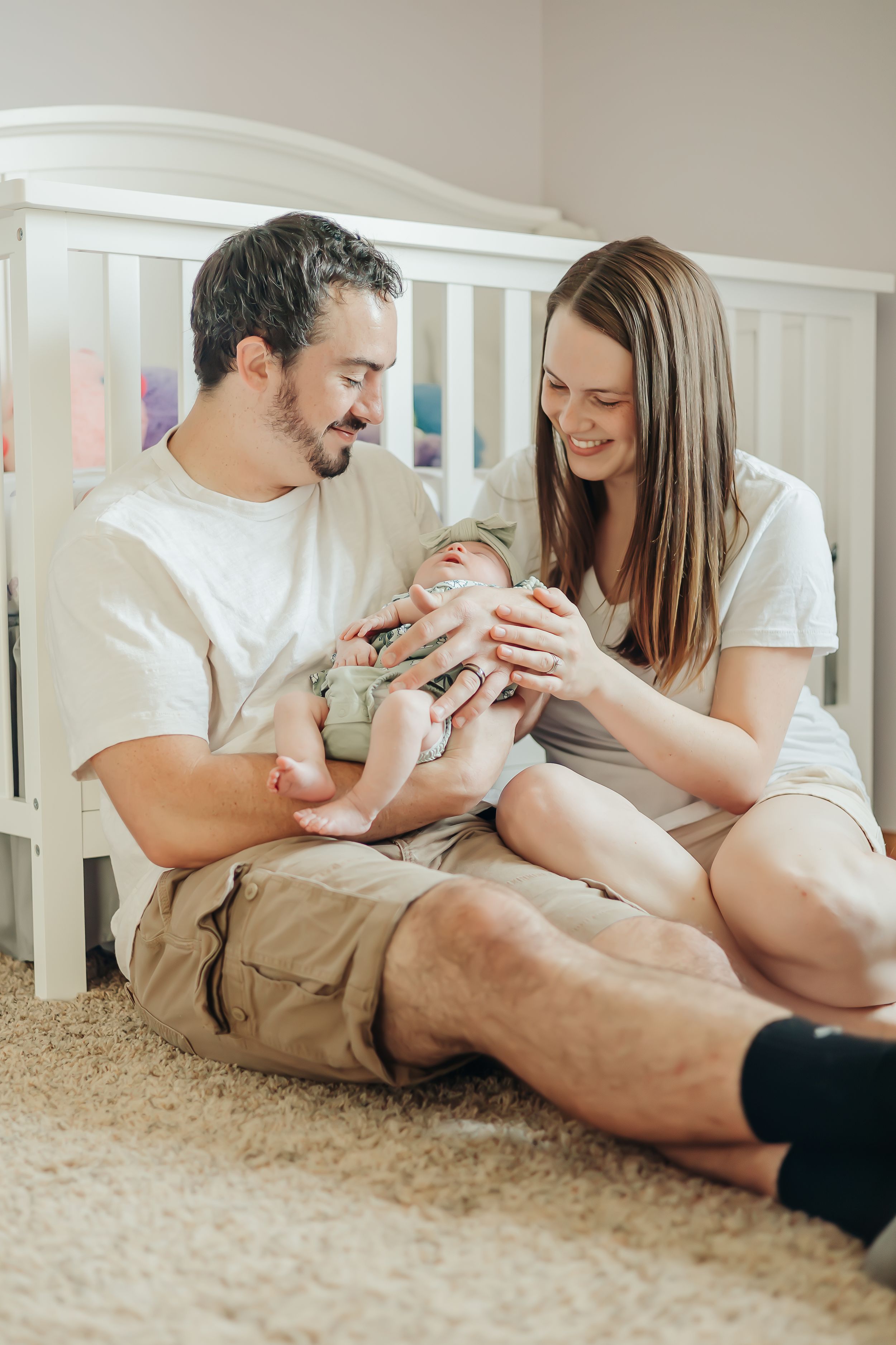 Parents sitting on carpet beside white crib with their newborn baby.