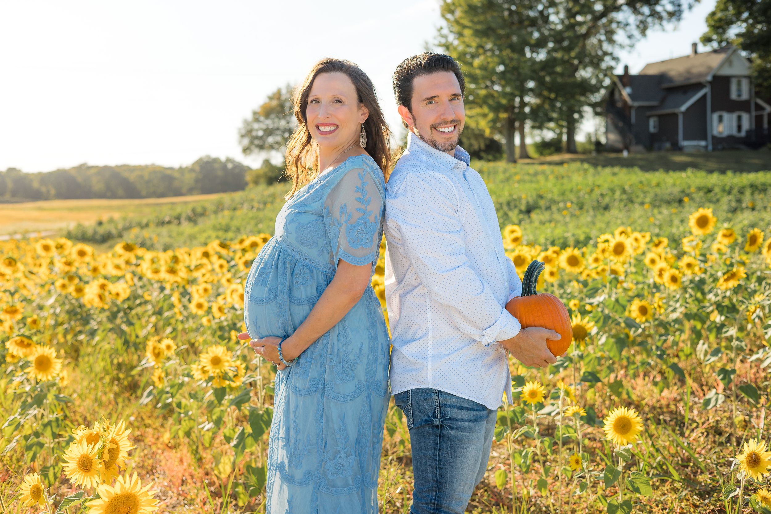 A couple stands back to back in a vibrant sunflower field during golden hour with a farmhouse in the background.