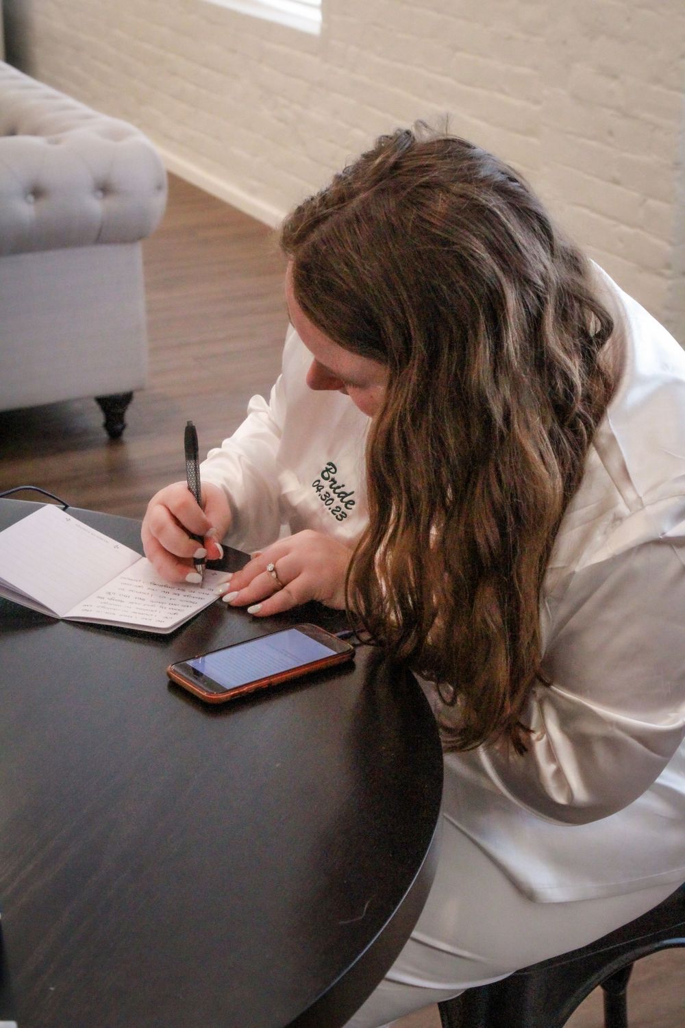 Someone writing in a notebook while referencing their phone, wearing a white sweater at a dark wooden table.