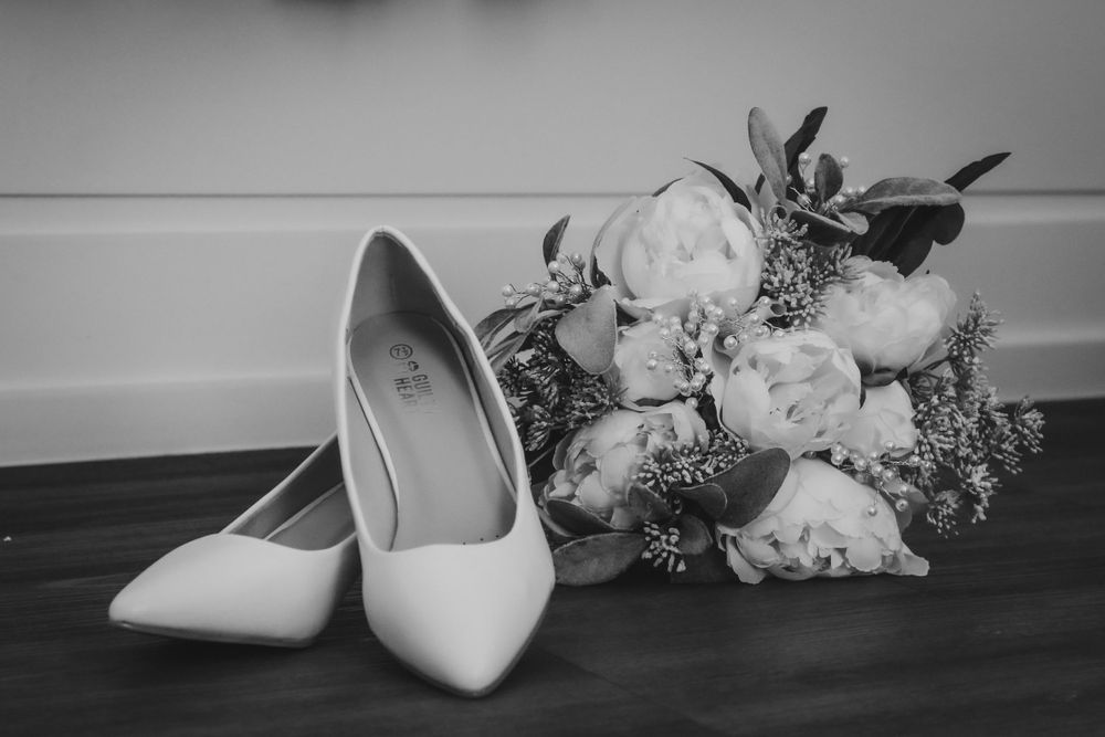 Elegant white bridal shoes next to a peony bouquet on dark wooden floor.