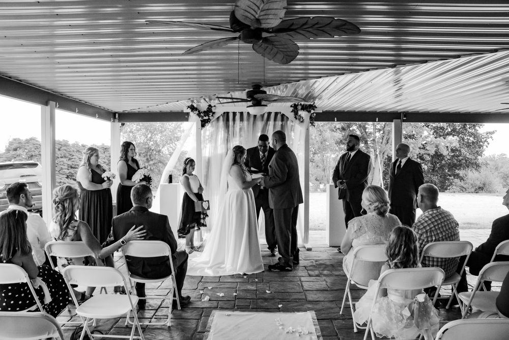 Wedding ceremony under covered porch with guests seated in white chairs.