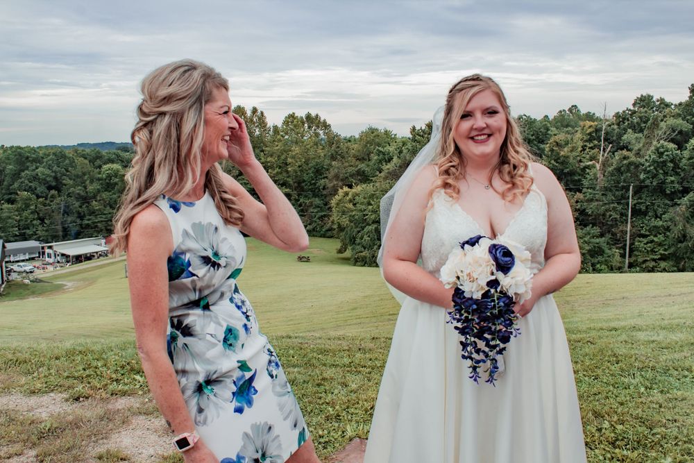 A candid outdoor wedding moment showing emotional reactions on a grassy hillside with trees in background.