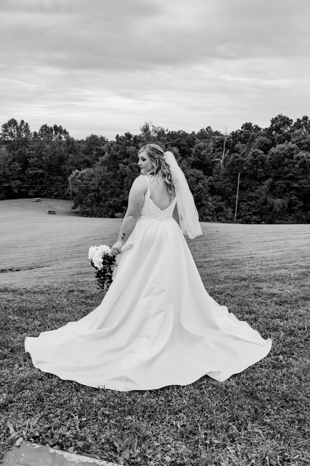 A dramatic black and white bridal portrait showing the full sweep of a wedding gown on a hilltop.