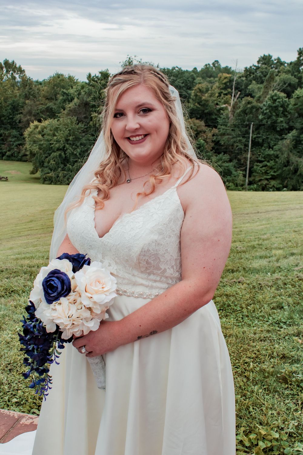 A radiant bride poses in a lace white gown holding a blue and peach bouquet against a scenic outdoor backdrop.