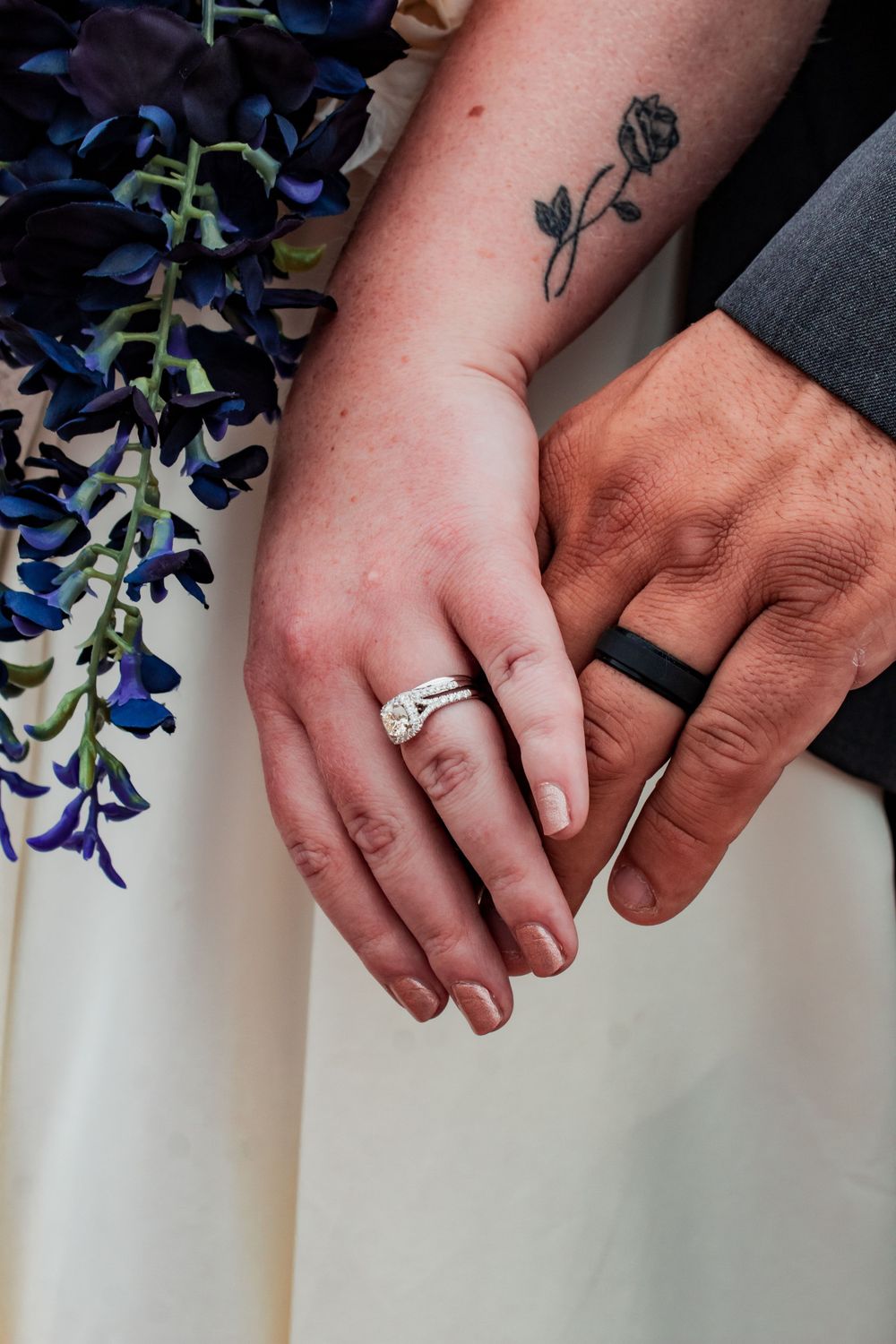 Close-up of newlyweds' hands showing wedding rings and rose tattoo with blue flowers in background.