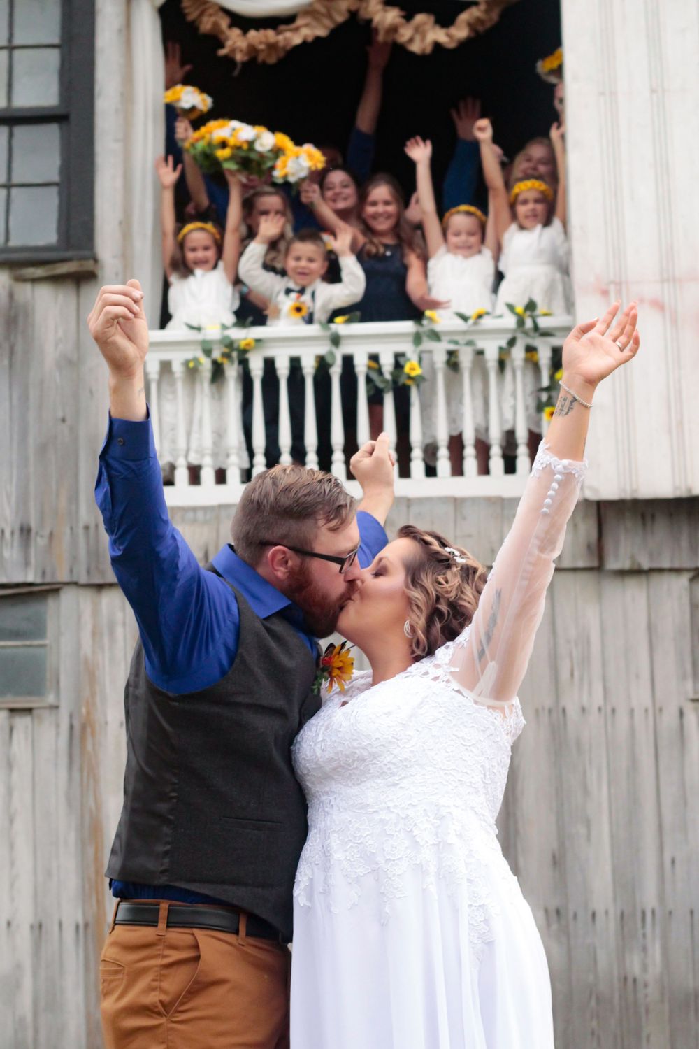 A celebratory wedding kiss with guests cheering from a white balcony decorated with yellow flowers.