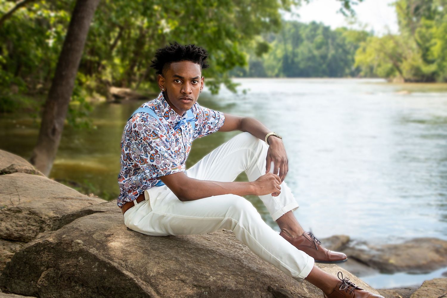 A person in a floral shirt poses casually on rocks by a peaceful river.