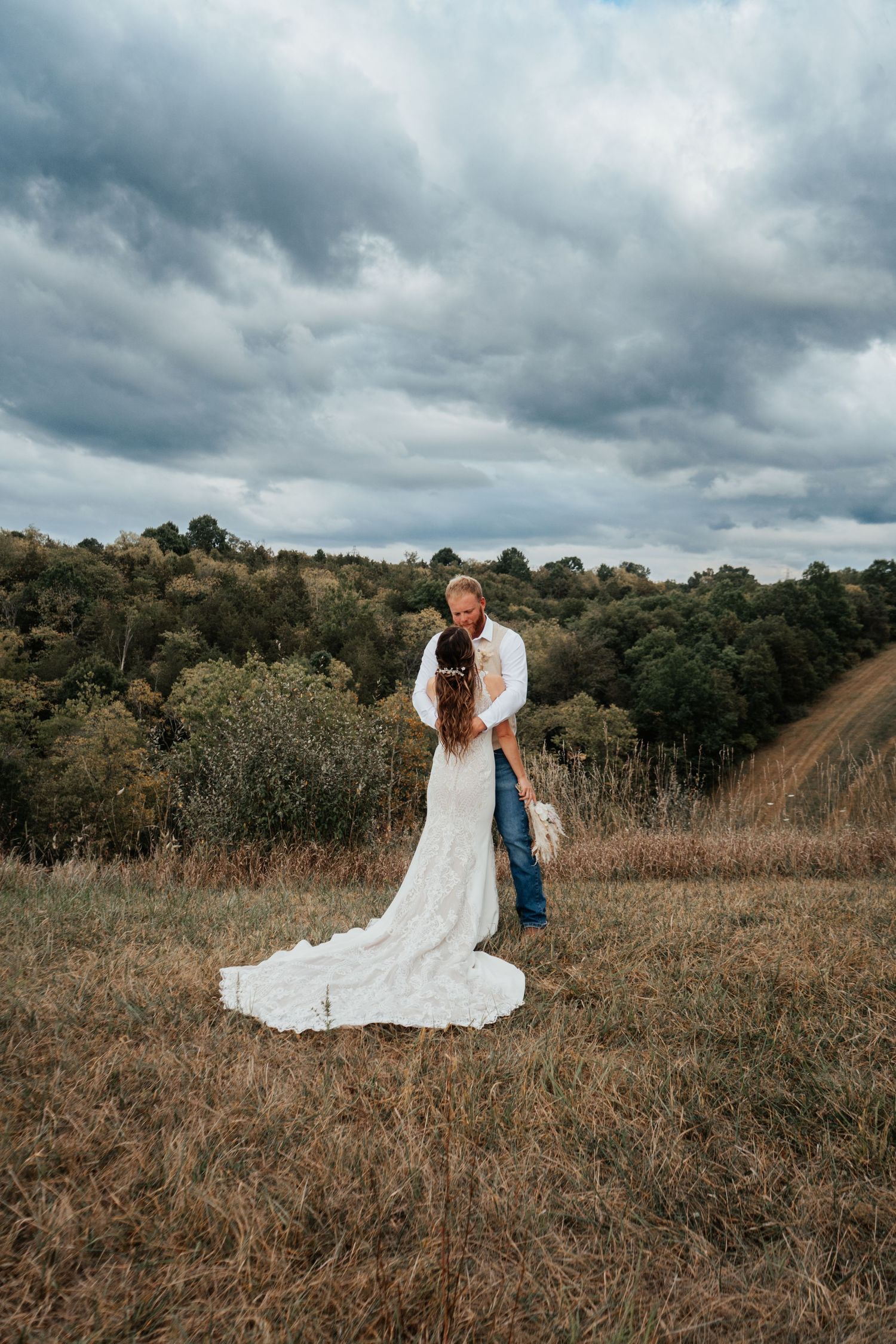 A couple in wedding attire poses in a rustic field beneath dramatic cloudy skies.