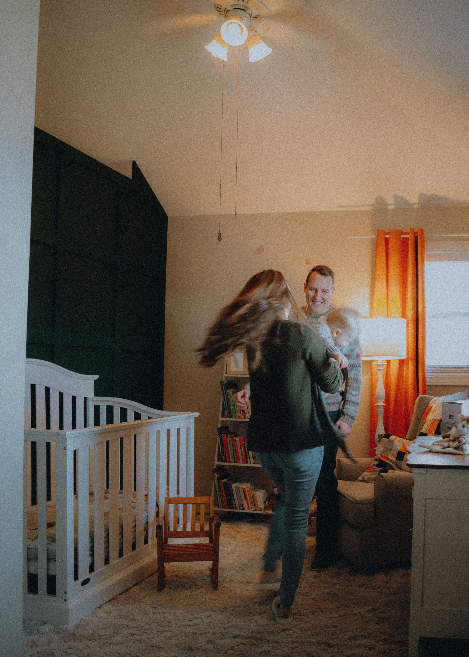 A home interior scene with a white crib, orange curtains, and a rocking chair in a warm-lit nursery.
