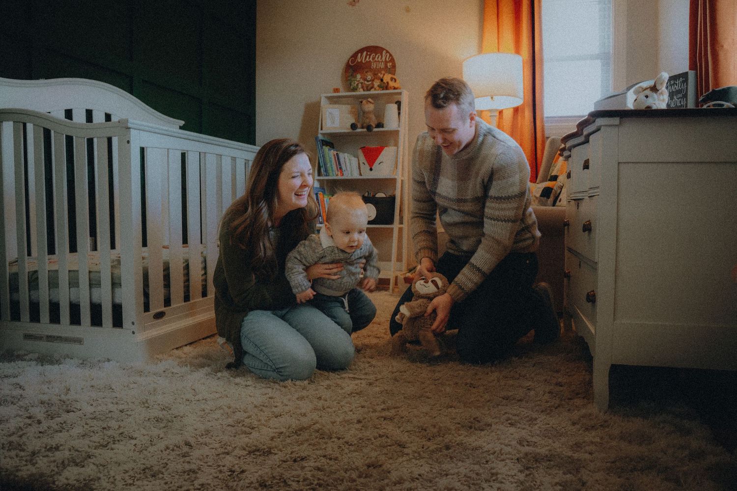 A cozy nursery scene with a family playing together on a carpeted floor beside a white crib and orange curtains in soft lighting.