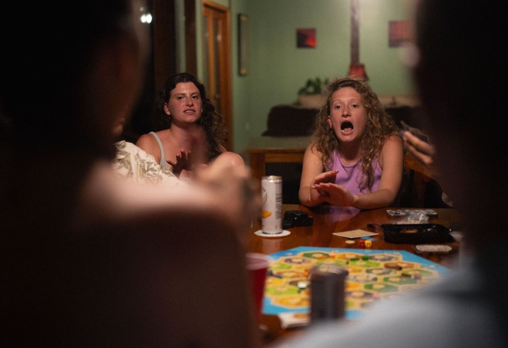 Board game pieces scattered across a table as players interact in low lighting.