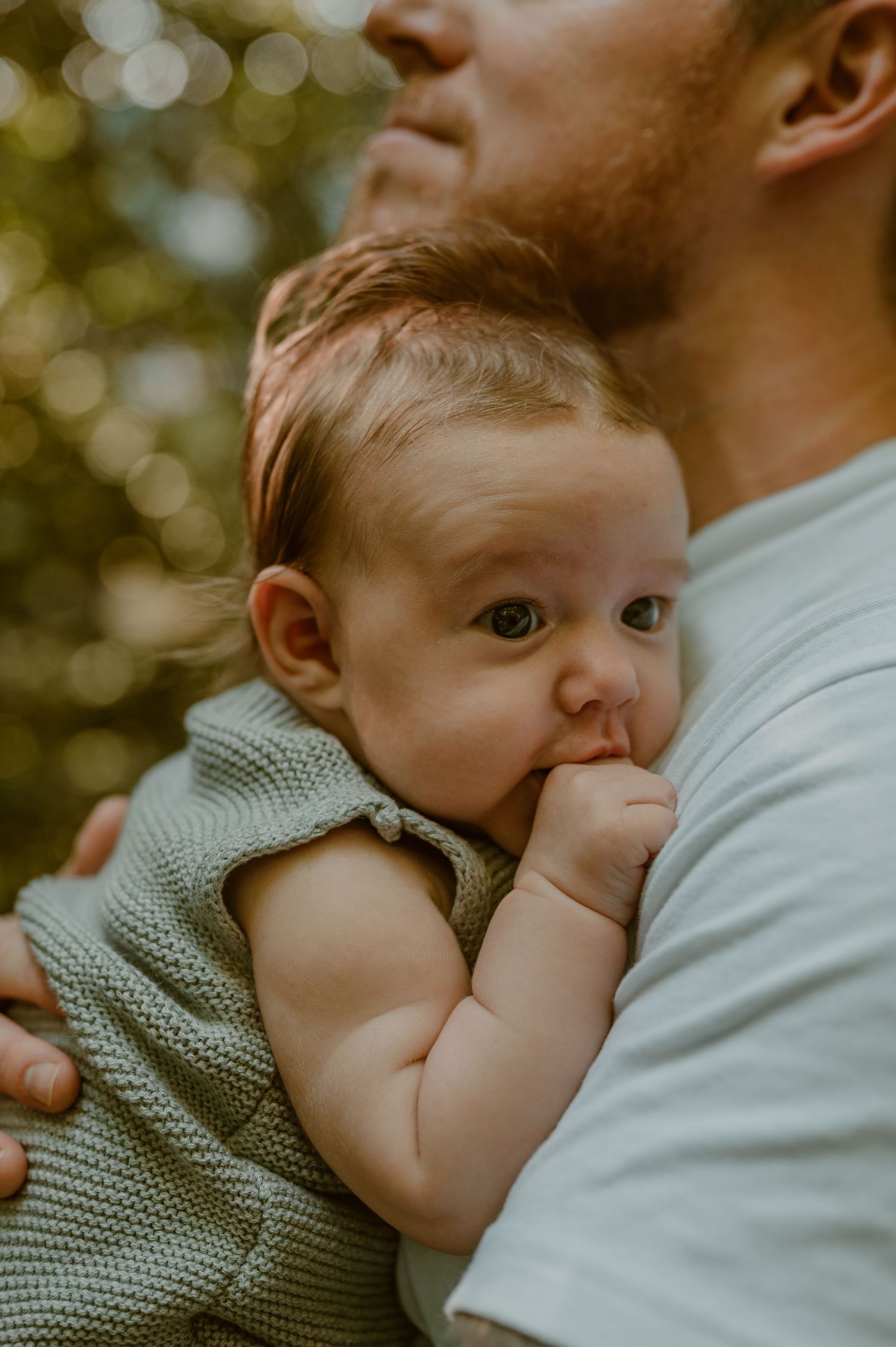 An infant wearing a knit mint green sweater nestles against someone's chest while sucking their thumb outdoors. photo by Samantha Campbell i