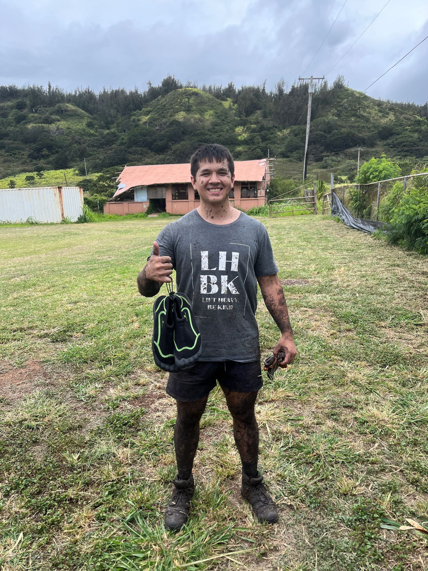 Person standing in a grassy field with farm buildings and mountains in the background.