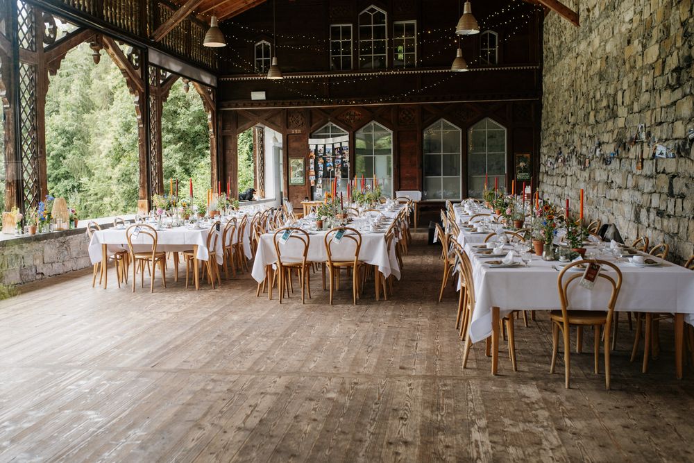 Rustic barn wedding reception with long white-draped tables, wooden chairs, string lights, and large arched windows.