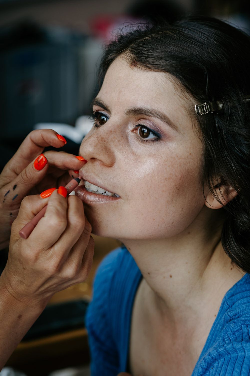 Makeup artist with red nails applying lipstick to a young brunette bride with freckles during wedding preparations.