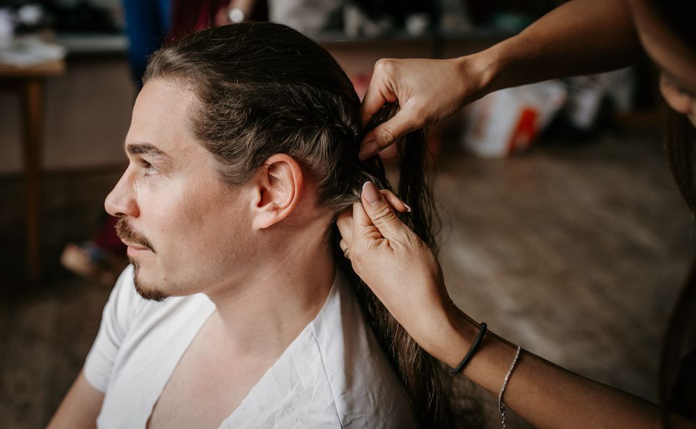 Hair stylist braiding long hair of a man in a white t-shirt during wedding preparation getting ready session.