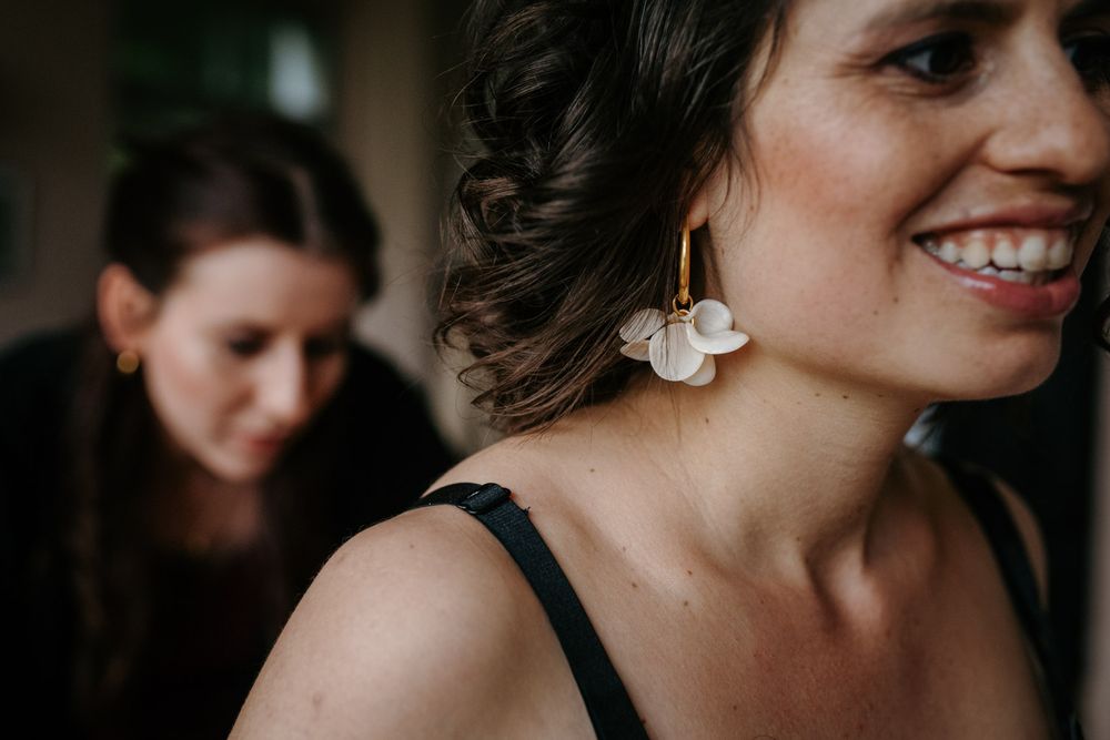 Smiling woman wearing gold and white floral earrings with a blurred bridesmaid in the background.