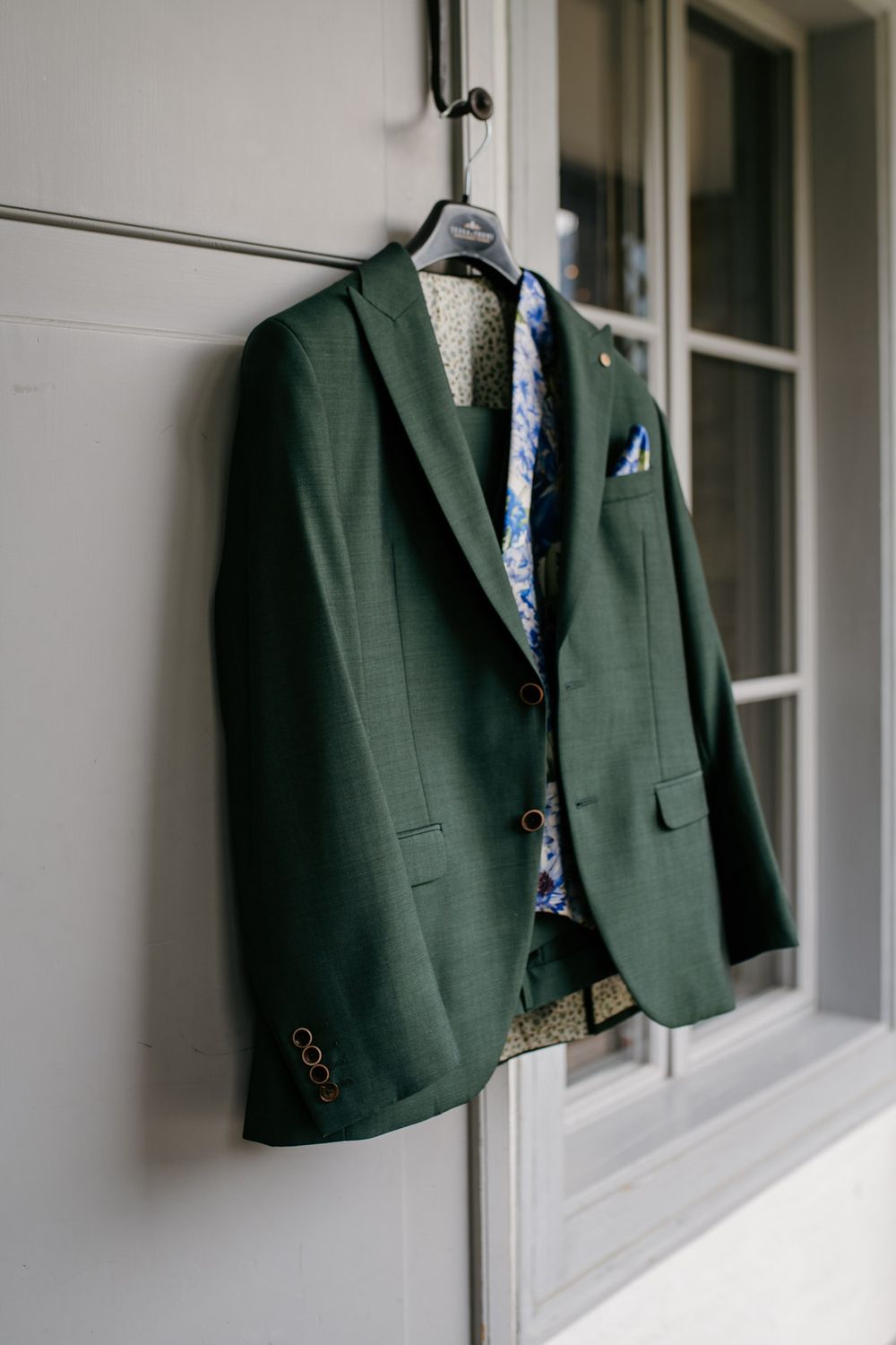 Dark green groom suit jacket with blue floral tie hanging on wall hook.