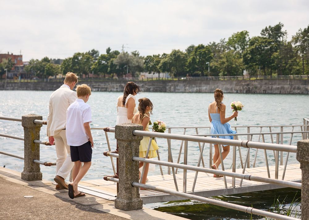 Wedding party walks along a waterfront dock, with bridesmaids in blue and yellow dresses holding bouquets on a sunny day.