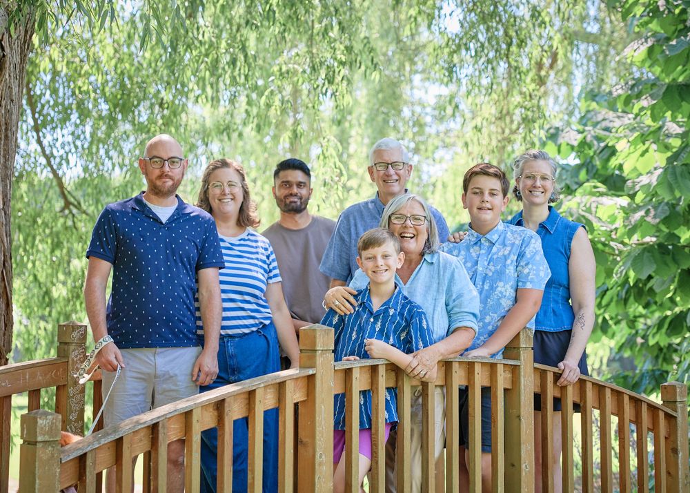 Multi-generational family of eight smiling together on a wooden bridge surrounded by lush green trees outdoors.