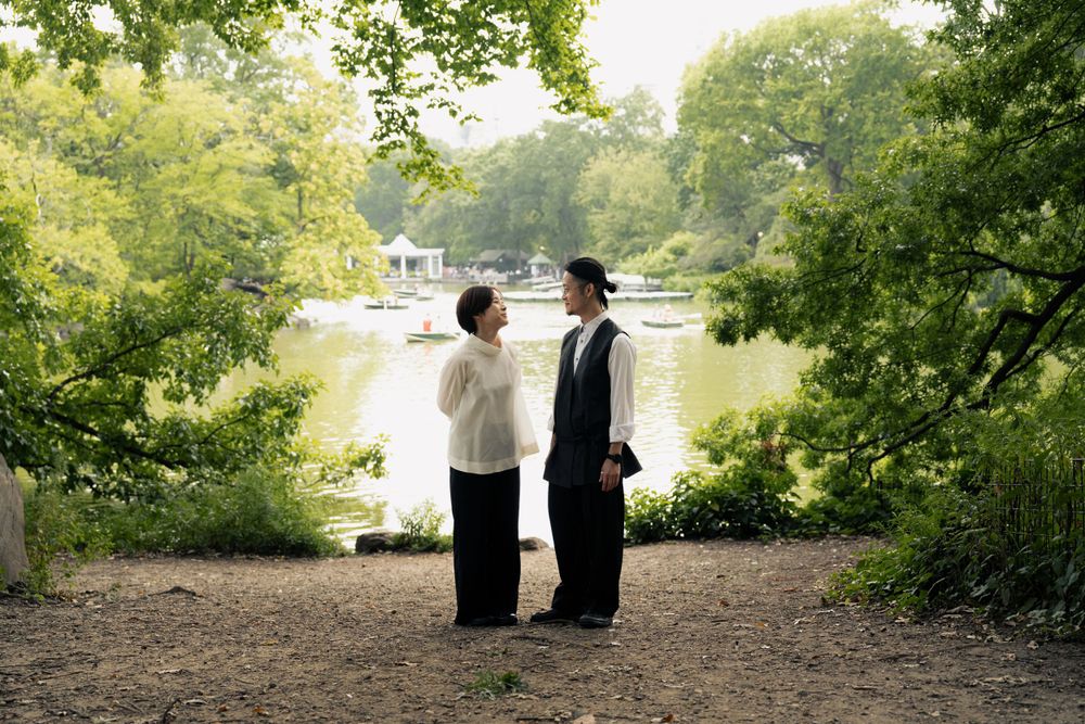 Couple in formal attire standing together under lush green trees in a romantic outdoor setting.